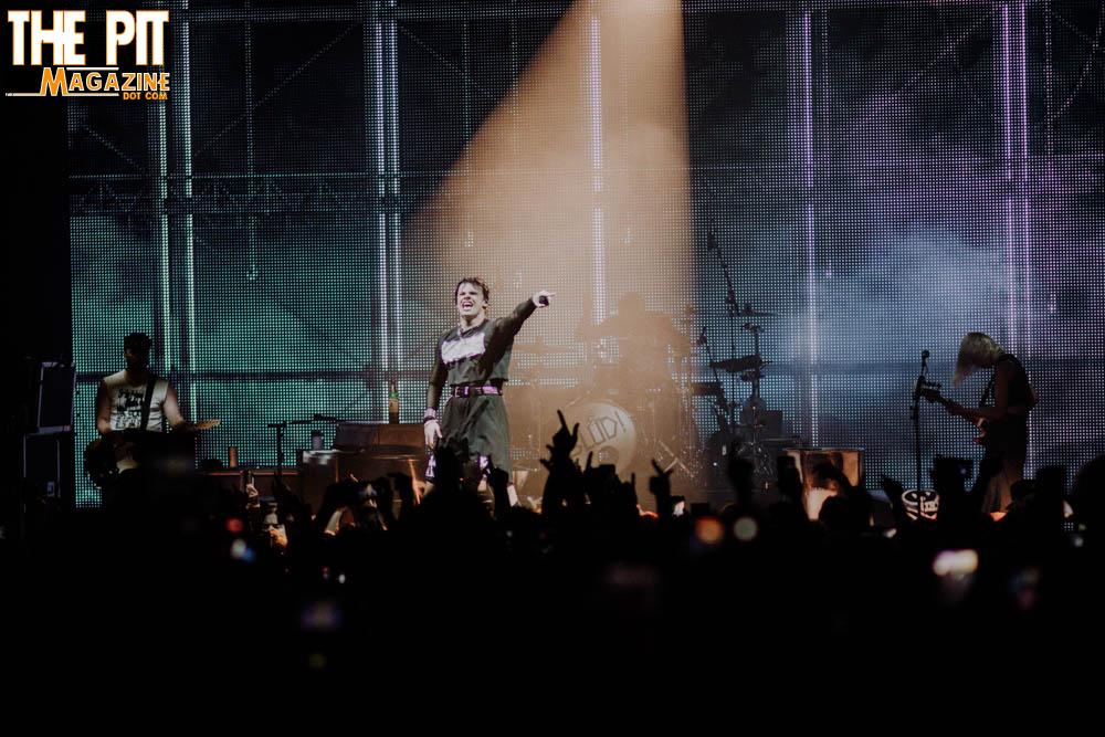 A rock band performing on stage, with Yungblud raising an arm triumphantly under bright stage lights while the audience watches.